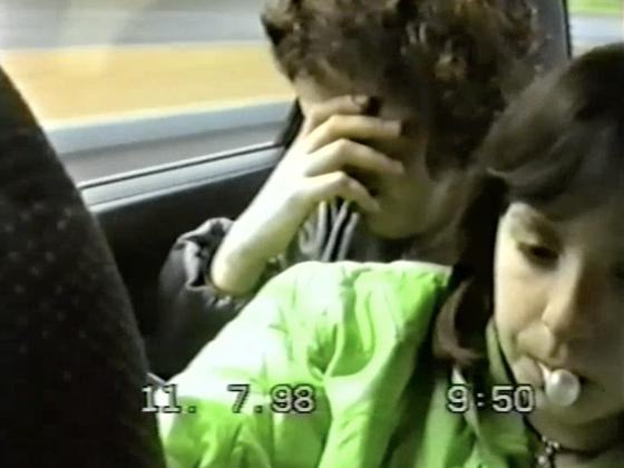 Filmstill: Zwei Kinder sitzen nebeneinander in einem Auto. Das rechte Kind hat eine aufgeblasene Kaugummiblase im Mund, das linke Kind hält sich die Hand vor das Gesicht.