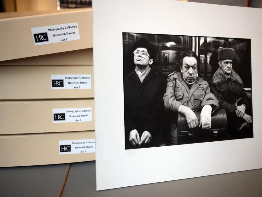Das Bild zeigt eine Fotografie Harald Hauswalds von drei sitzenden Männern in einer U-Bahn. Die Fotografie ist gegen einen Stapel Archivschachteln gelehnt.